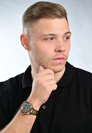 Gold wristwatch with a black face, metal bracelet, and a rotating bezel. Model wearing a black polo shirt, hand on chin, neutral background.