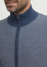 Sudadera azul con cremallera y cuello acanalado alto, que presenta un patrón de punto texturizado con franjas alternas claras y oscuras en tonos de azul.