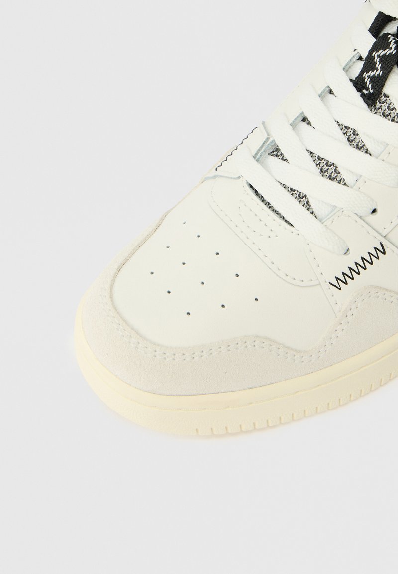 White sneaker featuring a suede toe cap and textured black laces. Perforated toe design, rubber sole with subtle tread pattern.