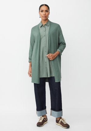 Cardigan verde aperto su una camicia button-up verde chiaro, abbinato a jeans in denim scuro con orli arrotolati e scarpe sportive marroni.