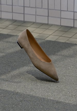 Chaussure plate beige à bout pointu flottant au-dessus d’une surface en asphalte grise avec un mur carrelé gris en arrière-plan.
