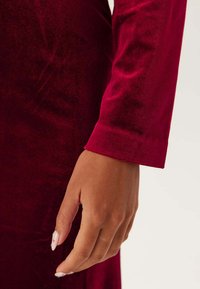 Röd sammetsklänning med långa ärmar. Närbild av �ärmdetalj som visar den släta texturen och den åtsittande manschetten. Nagellack synligt på handen.