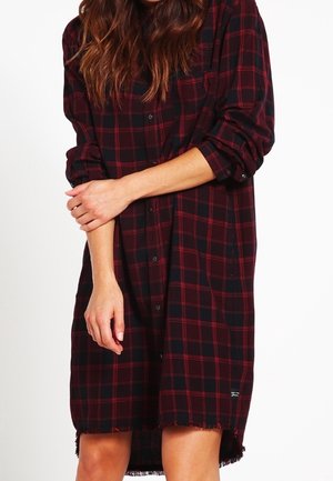 Ein rot-schwarzes Karo-Hemdkleid aus Stoff mit langen Ärmeln, Knopfleiste, fransigem Saum und lockerer Passform.