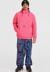 Νέος άνδρας που φοράει ένα φωτεινό ροζ μπουφάν με κουκούλα, μαύρο καπέλο, πολύχρωμο παντελόνι με σχέδια σε μπλε και ροζ και μαύρα παπούτσια, στέκεται με το πρόσωπο στραμμένο προς τα εμπρός.