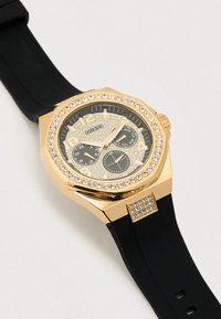 Orologio color oro con cinturino in silicone nero, decorato con cristalli intorno alla lunetta e quadrante nero testurizzato con più quadranti secondari.