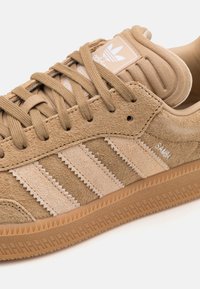 Tan suede sneaker with three beige stripes, textured finish, rubber outsole, and logo on tongue. Features rounded toe and flat laces.