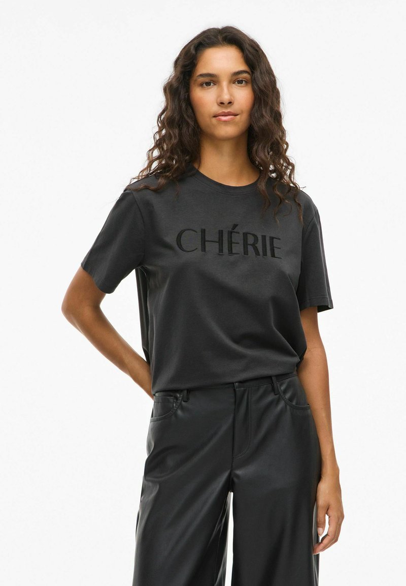 Sort bomulds T-shirt med hævet tekst "CHÉRIE" hen over brystet, parret med sorte bukser i kunstlæder. Korte ærmer og afslappet pasform.