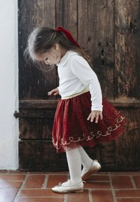 Gonna rossa glitterata con motivo a cuore sull'orlo, maglietta bianca a maniche lunghe, collant color crema e ballerine dorate lucide. Presenta un fiocco rosso per capelli.