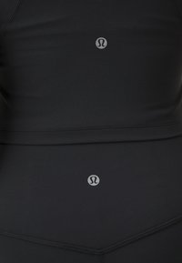 Top deportivo negro con un diseño ajustado, confeccionado en una tela elástica. Cuenta con un logo en el centro y detalles de costura visibles.