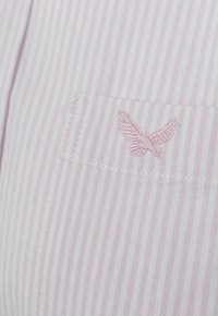 Camisa a rayas de color rosa claro con un bolsillo que presenta un diseño de ala bordado. La tela es suave y tiene un acabado fino a lo largo de las costuras.