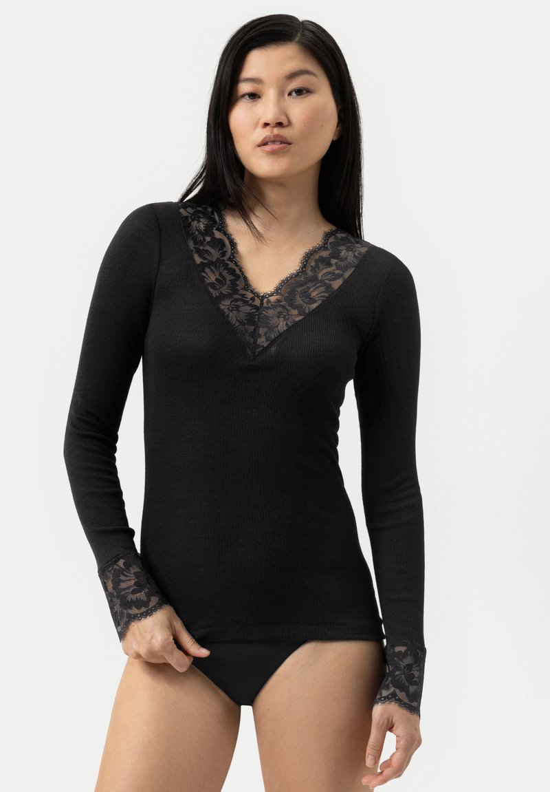 Kvinde iført en sort langærmet bluse med blomster-aldmissekant ved V-udskæringen og manchetterne, parret med sort undertøj, der står mod en hvid baggrund.