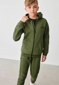 Olive zelen sweatshirt in usklajene trenirke iz mehke tkanine. Svetilka ima zadrgo, žepke in potisk na prsih. Obrobljena z žametnimi manšeti.
