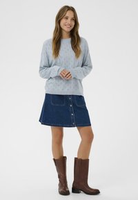 Pull en tricot bleu clair avec motif ajouré, assorti à une jupe en denim foncé avec des boutons sur le devant et deux poches avant. Bottes brunes jusqu'aux genoux.