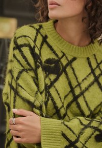 Green knitted sweater featuring a black geometric pattern, ribbed neckline, and cuffs. The fabric has a soft, fluffy texture.