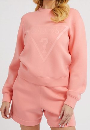 Kobieta ubrana w dopasowany różowy komplet z bluzą i szortami, z wytłoczonym logo Guess na przodzie bluzy.