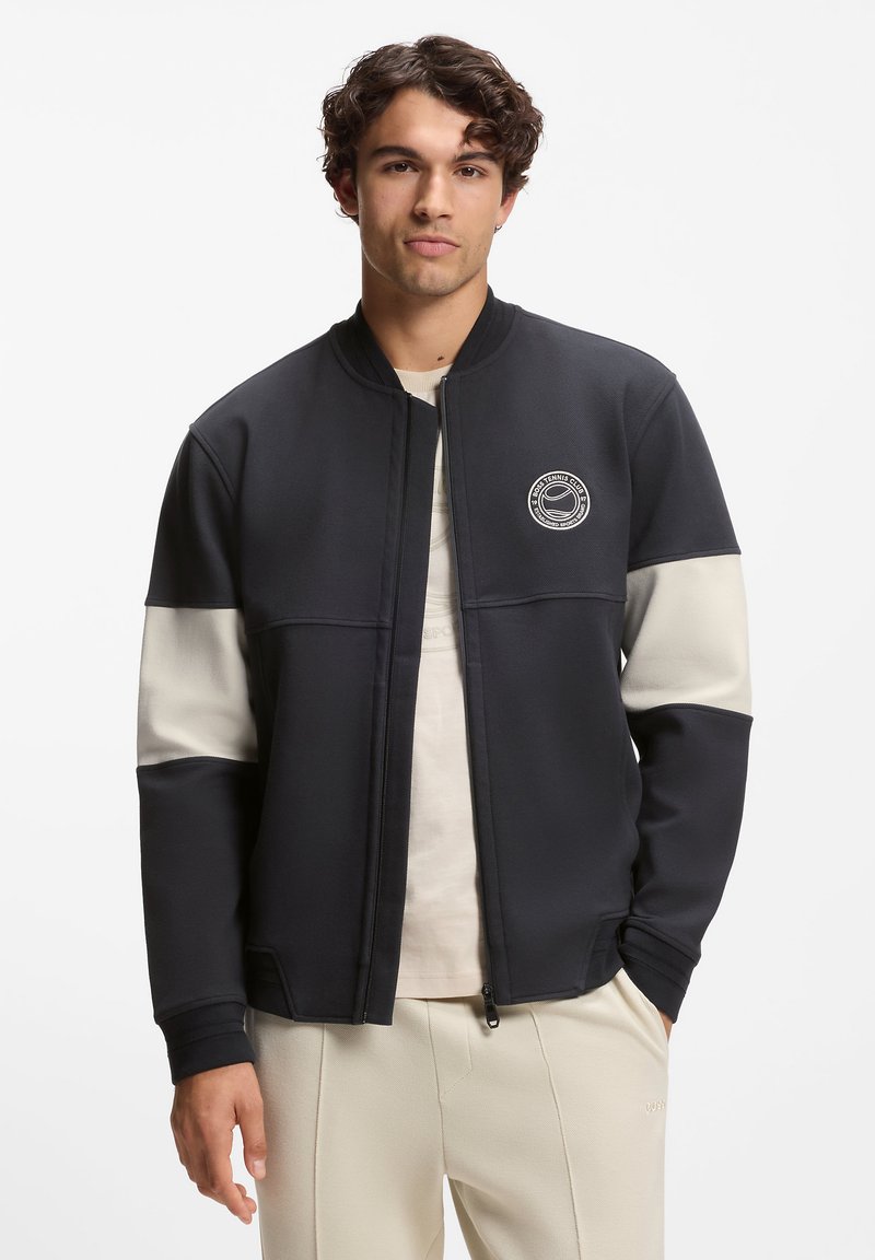 Schwarze und cremefarbene Sportjacke mit frontalem Reißverschluss, gerippten Bündchen und rundem Logo auf der Brust. Glattes Gewebe, figurbetontes Design.