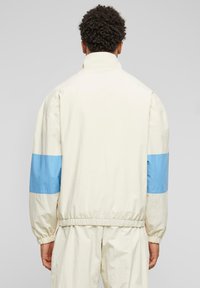 White lightweight jacket with blue accents on sleeves and elastic cuffs. Features a high collar and a relaxed fit design.