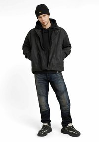 Black leather jacket over a black hoodie, blue tapered jeans with stitch detailing, and black sneakers with gray accents.