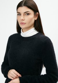 Schwarzer, flauschiger Pullover mit langen Ärmeln und rundem Ausschnitt, über einem weißen Rollkragenpullover getragen. Weiche Textur und minimalistisches Design.
