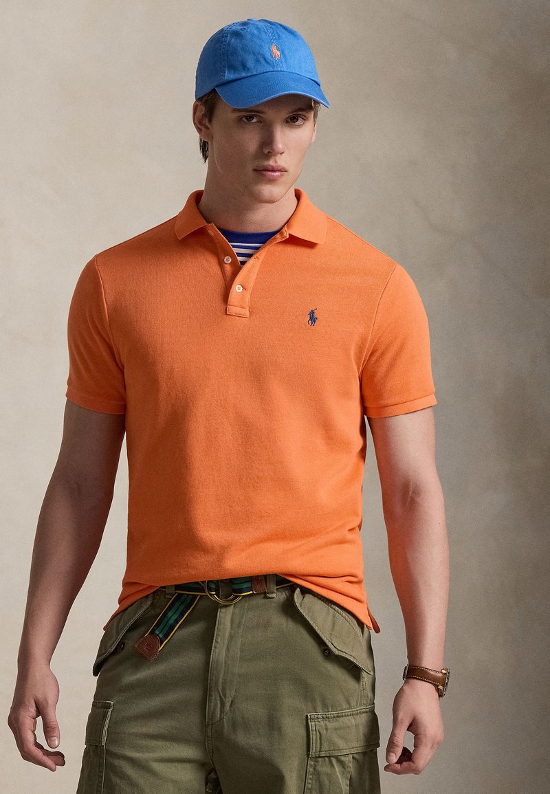 Νέος άνδρας φορώντας πορτοκαλί πόλο πουκάμισο, μπλε καπέλο μπέιζμπολ, ελιά παντελόνια cargo με ριγωτή ζώνη και καφέ ρολόι χειρός σε ουδέτερο φόντο.