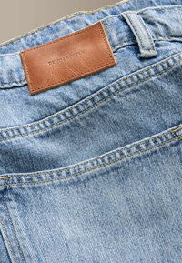 Poche arrière d'un jean en denim bleu clair avec un patch en cuir marron cousu portant la mention "DENIM SUPPLY" à la taille.