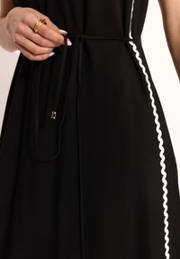 Czarna sukienka z wiązaniem w talii i białymi, falbaniastymi wykończeniami. Gładki materiał, podkreślający smukłą sylwetkę i minimalistyczne detale projektu.