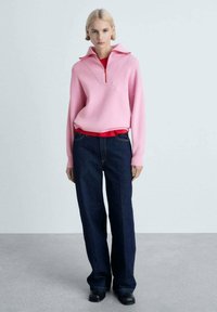 Rosa Strickpullover mit geripptem Kragen und Reißverschluss, über einem roten Hemd getragen, kombiniert mit dunkelblauen weit geschnittenen Jeans und schwarzen Schuhen.