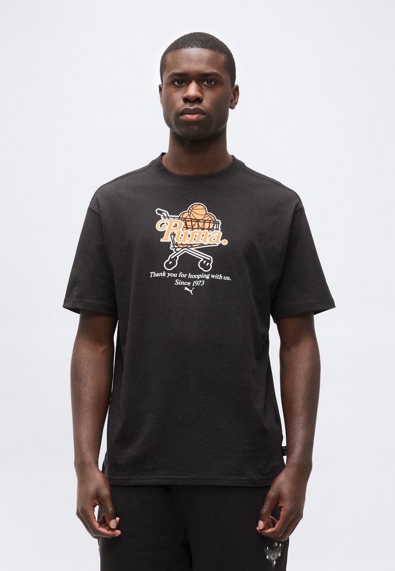 Άνδρας που φοράει μαύρο μπλουζάκι με γραφικό καρότσι αγορών, κρατώντας μπάλες μπάσκετ και κείμενο «Puma. Σας ευχαριστούμε που παίζετε μαζί μας. Από το 1973.»