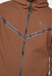 Braune Zip-Up-Hoodie aus weichem Stoff, mit einem grauen Streifenmuster und einem Nike-Logo auf der Brust. Mit Reißverschluss und Kapuze.