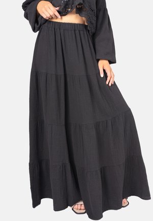 Frau trägt einen langen, gestuften schwarzen Rock mit geraffter Taille und ein schwarzes, kurz geschnittenes Oberteil mit weiten Ärmeln, hält den Rocksaum mit einer Hand.