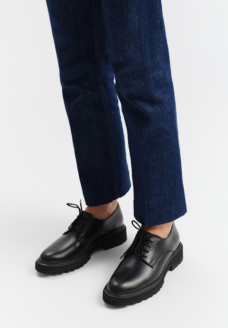 Czarne skórzane buty wizytowe z okrągłymi noskami, niską podeszwą oraz cienkimi sznurowadłami, w połączeniu z ciemnoniebieskimi dżinsami. Odpowiednie dopasowanie w okolicy kostki.
