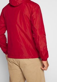 Veste légère rouge avec capuche, poignets élastiques et texture lisse. Associée à un pantalon beige, montrant un design simple et fonctionnel.