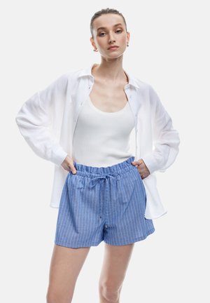 Chemise blanche à manches longues portée par-dessus un débardeur blanc, associée à un short bleu à rayures avec une taille élastique et un cordon de serrage.