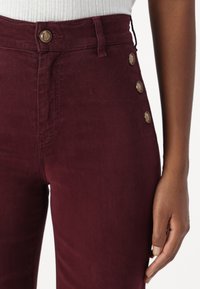 Pantaloni bordeaux a vita alta in tessuto liscio, con chiusura frontale a bottone e bottoni decorativi laterali per un tocco di stile.