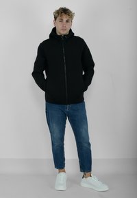 Felpa nera con zip e cappuccio, tasche laterali e tessuto testurizzato, abbinata a jeans slim fit azzurri e sneakers bianche.