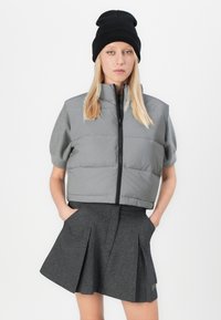 Donna con un berretto nero, gilet imbottito grigio a maniche corte con zip e una gonna plissettata grigio scuro, con le mani nelle tasche, su sfondo bianco.