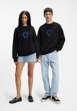 Kobieta i mężczyzna stoją obok siebie, ubrani w czarne bluzy z niebieskim logo uśmiechu; kobieta w dżinsowej spódnicy i mokasynach, mężczyzna w jasnych dżinsach i trampkach.