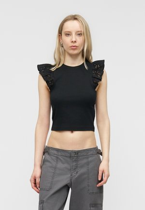 Hosszú szőke hajú nő fekete ujjatlan felsőben, amelyen csipkés fodros vállrész található, és szürke cargo nadrágban, világos háttér előtt áll.