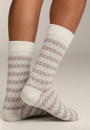 Gambe con calze di lana crema con modelli a zigzag di un marrone chiaro, un piede piatto e l'altro leggermente sollevato e incrociato dietro.