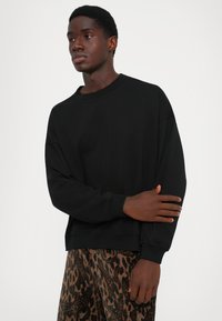 Mladý muž v čiernom oversized mikine a hnedých vzorovaných nohaviciach, stojí s jednou rukou presehnutou cez telo na jednoduchom pozadí.