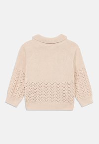 Vaaleanbeige neulottu neulepusero, jossa on korkea kaulus, alareunassa on suihkulähdekuvio ja resorihihansuut.