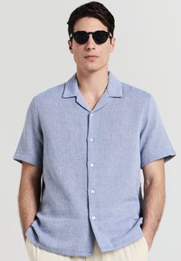 Uomo che indossa occhiali da sole neri a forma tonda e una camicia azzurro chiaro a maniche corte con un motivo tessuto, con le mani nelle tasche dei pantaloni beige.