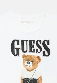 Λευκό βαμβακερό t-shirt με γραφικό αρκούδας σε αποχρώσεις του καφέ, που φοράει πουκάμισο και κρατά τσάντα· η λέξη "GUESS" τυπωμένη με έντονα μαύρα γράμματα.