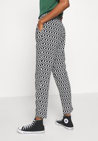 Pantalon à motif noir et blanc, coupe slim avec poches, présentant un design hexagonal et une ceinture élastique, assorti à des chaussures montantes noires.
