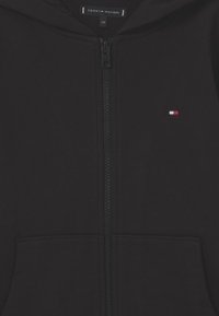 Schwarze Reißverschluss-Hoodie aus weicher Baumwolle. Verfügt über einen Frontreißverschluss, zwei Taschen und ein kleines Logo in Rot, Weiß und Blau auf der linken Brust.