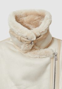 Abbigliamento esterno color crema con collare in pelliccia sintetica. Presenta una chiusura con zip e una fascia in pelle con fibbia. Superficie testurizzata.