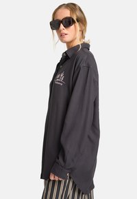 Camisa de manga larga gris oscuro con cuello, que presenta un bordado de un cactus rosa y detalles de botones, combinada con unas gafas de sol negras de gran tamaño.