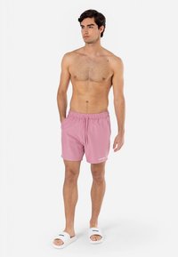 Rozā peldbikses ar elastīgu jostu un aukliņu. Balta logotipa detaļa sānos. Modelis valkā baltas čības, stāvot uz vienkārša fona.