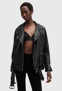 Black leather biker jacket featuring silver studs on shoulders, asymmetrical zip closure, and side pockets. Model wears a black bikini top.
