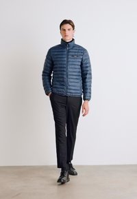 Giacca a vento blu con trapuntatura orizzontale, chiusura con zip, colletto alto, abbinata a pantaloni e scarpe neri, su uno sfondo semplice.